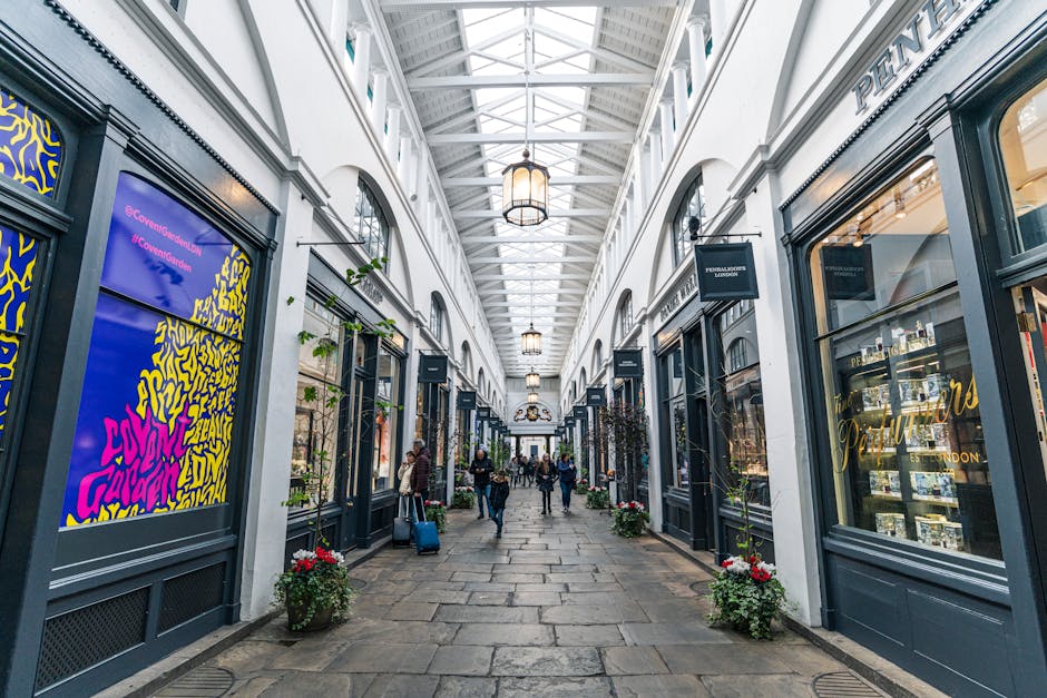 The image depicts a covered shopping arcade with a high, white, vaulted ceiling supported by metal framework, illuminated by hanging lantern-style light fixtures. Along both sides of the walkway, shop