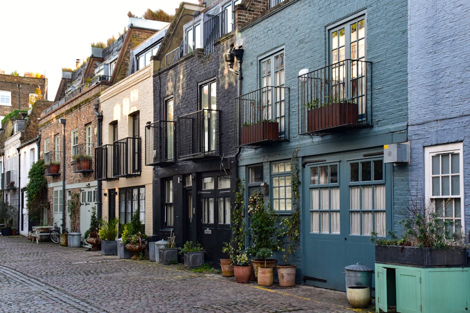 A row of terraced townhouses along a cobblestone street, each featuring small balconies with black metal railings and potted plants, including lush green foliage and flowering plants in clay and plast